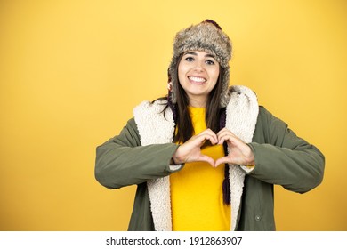 Young Beautiful Woman Wearing A Hat And A Green Winter Coat Over Yellow Background Smiling In Love Showing Heart Symbol And Shape With Hands