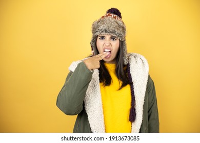 Young Beautiful Woman Wearing A Hat And A Green Winter Coat Over Yellow Background Disgusted With Her Hand Inside Her Mouth