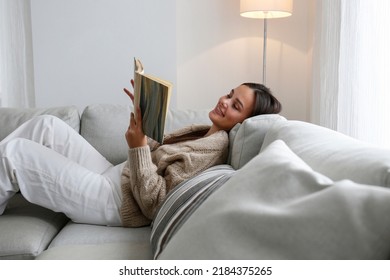 Young beautiful woman wearing knitted cardigan reading a book at home. Brunette female in a beige sweater sitting on textile couch. White wall background, copy space, close up. - Powered by Shutterstock