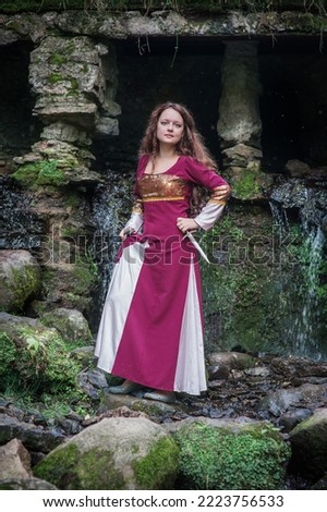 Young beautiful woman wearing medieval style dress with dagger near waterfall outdoor