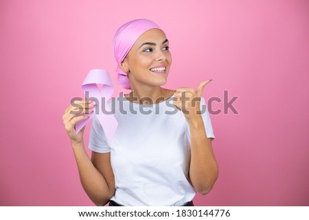 Young beautiful woman wearing pink headscarf holding brest cancer ribbon over isolated pink background smiling and pointing with thumb to the side