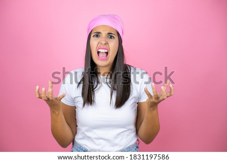 Young beautiful woman wearing pink headscarf over isolated pink background crazy and mad shouting and yelling with aggressive expression and arms raised. Frustration concept.