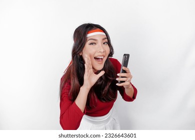 Young beautiful woman wearing a red top is holding her phone while shouting and screaming loud with a hand on her mouth. Indonesia's independence day concept.