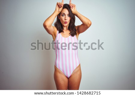 Young beautiful woman wearing striped pink swimsuit swimwear over isolated background doing funny gesture with finger over head as bull horns