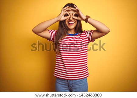 Young beautiful woman wearing striped t-shirt standing over isolated yellow background doing ok gesture like binoculars sticking tongue out, eyes looking through fingers. Crazy expression.