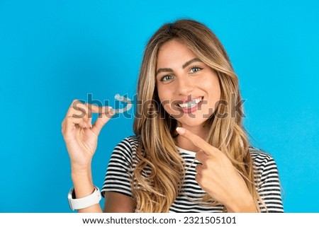 Young beautiful woman wearing striped t-shirt holding an invisible aligner and pointing at it. Dental healthcare and confidence concept.