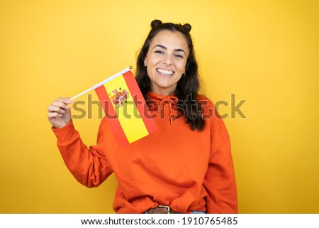 Young beautiful woman wearing sweatshirt over isolated yellow background holding flag of Spain with a happy face standing and smiling with a confident smile showing teeth