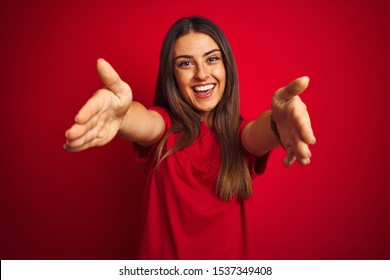 Young beautiful woman wearing t-shirt standing over isolated red background looking at the camera smiling with open arms for hug. Cheerful expression embracing happiness.