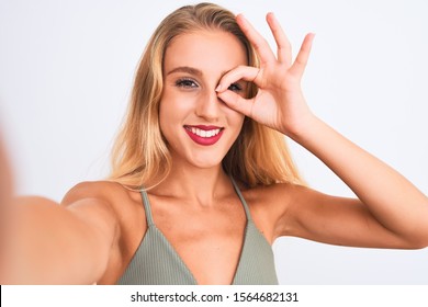 Young beautiful woman wearing t-shirt make selfie by camera over isolated white background with happy face smiling doing ok sign with hand on eye looking through fingers - Shutterstock ID 1564682131