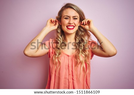 Young beautiful woman wearing t-shirt standing over pink isolated background Smiling pulling ears with fingers, funny gesture. Audition problem