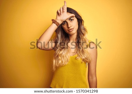 Young beautiful woman wearing t-shirt over yellow isolated background making fun of people with fingers on forehead doing loser gesture mocking and insulting.