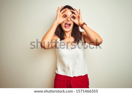 Young beautiful woman wearing t-shirt standing over white isolated background doing ok gesture like binoculars sticking tongue out, eyes looking through fingers. Crazy expression.