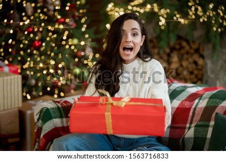 Young beautiful woman wearing warm sweater holding present sitting on the sofa in room decorated for celebrating new year and christmas looking surprised festive mood