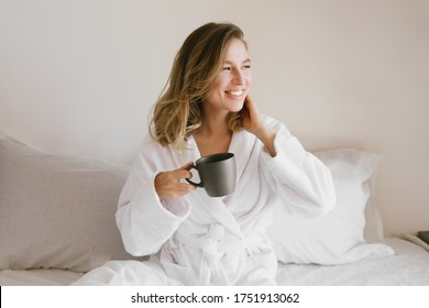 Young beautiful woman wearing white bathrobe having breakfast in bed with coffee and croissant and fresh fruits in cozy bedroom. Morning rituals.