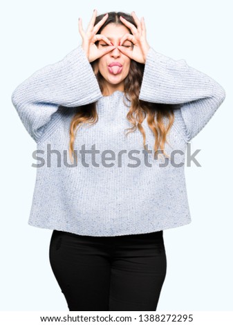 Young beautiful woman wearing winter sweater doing ok gesture like binoculars sticking tongue out, eyes looking through fingers. Crazy expression.