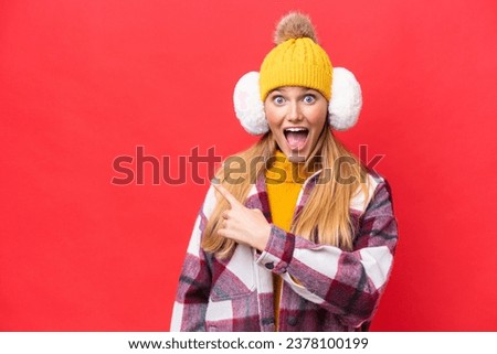 Young beautiful woman wearing winter muffs isolated on red background surprised and pointing side
