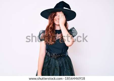 Young beautiful woman wearing witch halloween costume covering one eye with hand, confident smile on face and surprise emotion. 