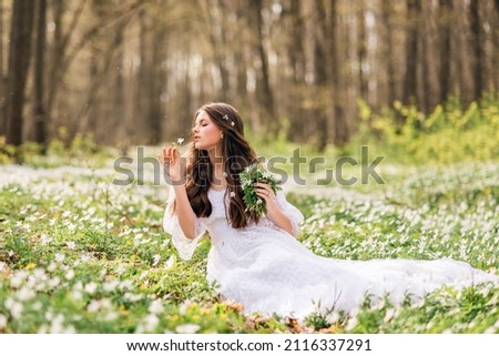 young beautiful woman in a white long dress collects primroses. Fairy girl in the spring forest. A bouquet of white flowers in her hands