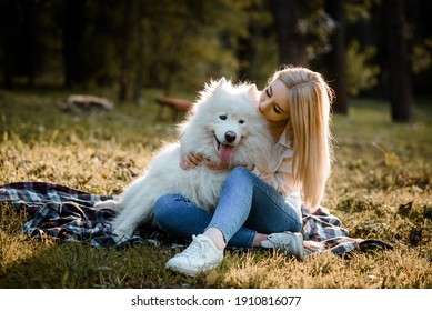 Young beautiful woman in white shirt is hugging and kissing her white dog samoyed outdoors in the park and sitting on the grass.