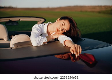 young beautiful woman in a white shirt sitting in a red car cabriolet with a white interior. red car in the field 
on a sunny day. the wind blows the girl's hair. red cabriolet. 