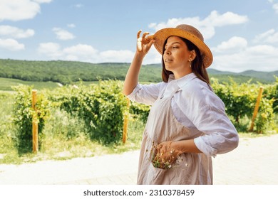 Young beautiful woman in white shirt, beige sundress and straw hat looks dreamily at sky. Attractive brunette smiles against backdrop of vineyards and green hills. Agriculture and village life concept - Shutterstock ID 2310378459