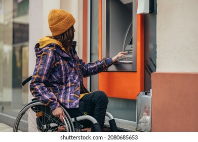 Young beautiful woman who uses a wheelchair using credit card and atm machine while withdrawing money
