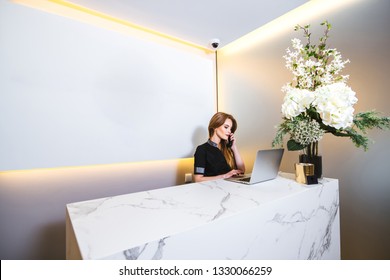 Young Beautiful Woman Working In The Modern Reception Of A Clinic Answering A Phone Call While Consulting The Computer To See Available Appointments