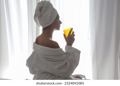 Young beautiful woman wrapped in towels drinking liquid collagen or vitamin supplement next to the window, healthy morning lifestyle concept - Shutterstock ID 2324041085