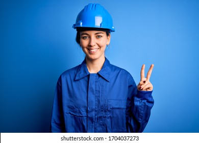 Young beautiful worker woman with blue eyes wearing security helmet and uniform smiling with happy face winking at the camera doing victory sign. Number two.