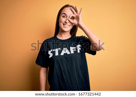Young beautiful worker woman wearing staff uniform t-shirt over isolated yellow background doing ok gesture with hand smiling, eye looking through fingers with happy face.