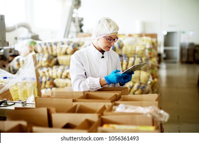 Young beautiful worried female worker in sterile clothes is using a tablet while being leaned against the stacks of open boxes inside of factory storage. - Powered by Shutterstock