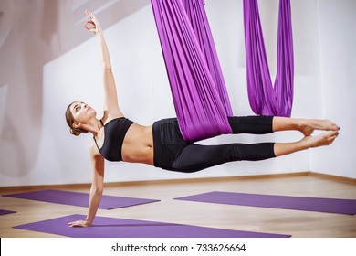 Young beautiful yogi woman doing aerial yoga practice in purple hammock in fitness club.