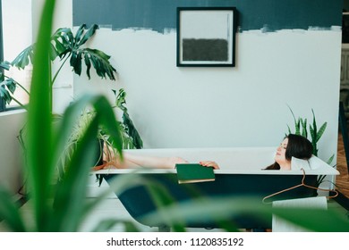 Young Beautifull Girl In Pink Dress Lying In Empty Vintage Cast-iron Bath Inside Decorative In French Style Room With Green Plants. Odd Unusual Strange Woman Home Relaxation. Comfortable Designer Bed