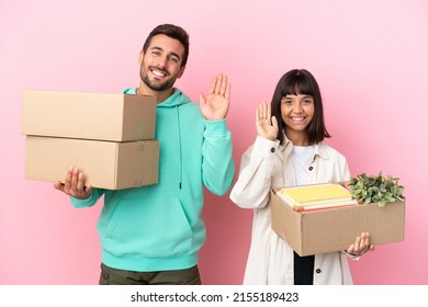 young beauty couple moving in new home among boxes isolated on pink background saluting with hand with happy expression