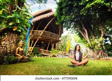 Young beauty sexy woman practices yoga in the tropical garden on a background scenic nature landscape with big green tree and beautiful bamboo house at sunny summer day