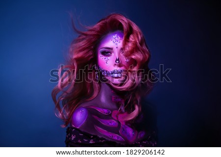 A young beauty woman with long hair in the image of a sugar skull. Hair flying in wind. Halloween carnival party. Close up portrait. A cute sad face in neon colored light