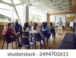 Young berded man speaking on a business training standing in office during a conference or seminar for a group of diverse people company employees sitting on chairs in meeting room.