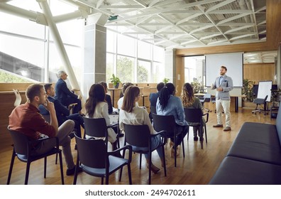 Young berded man speaking on a business training standing in office during a conference or seminar for a group of diverse people company employees sitting on chairs in meeting room. - Powered by Shutterstock