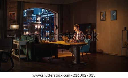 Young Beuatiful Women Working from Home, Sitting And Typing on Laptop Computer in Stylish Loft Apartment in the Evening. Creative Female Wearing Cozy Casual Outfit. Urban City View from Big Window.