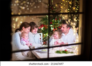 Young big family celebrating Christmas enjoying dinner, view from outside through a window into a decorated living room with tree and candle lights, happy parents eating with three kids