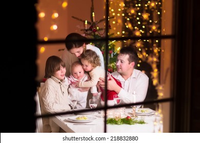 Young Big Family Celebrating Christmas Enjoying Dinner, View From Outside Through A Window Into A Decorated Living Room With Tree And Candle Lights, Happy Parents Eating With Three Kids