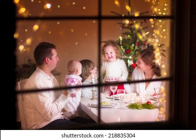 Young Big Family Celebrating Christmas Enjoying Dinner, View From Outside Through A Window Into A Decorated Living Room With Tree And Candle Lights, Happy Parents Eating With Three Kids 