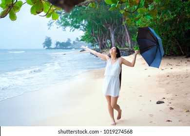 Young biracial Asian Caucasian woman or teen walking on beach holding umbrella on overcast rainy day, arms outstreched enjoying the rain, smiling - Powered by Shutterstock