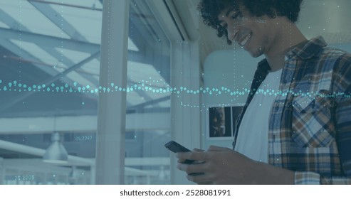 A young biracial chef with curly black hair and beard is looking at a smartphone. Wearing a plaid shirt, he stands by a window, smiling at screen - Powered by Shutterstock