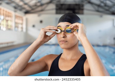 Young biracial female swimmer adjusting goggles at an indoor pool. She has short black hair, light brown skin, and is wearing a black swimsuit, unaltered. - Powered by Shutterstock