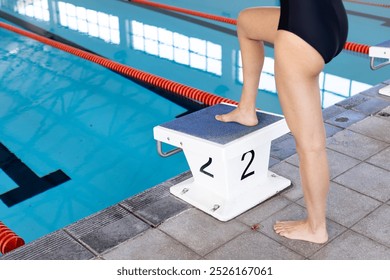 Young biracial female swimmer preparing to dive into an indoor pool, copy space. She has dark hair, wearing a black swimsuit, on starting block, unaltered - Powered by Shutterstock