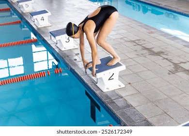 Young biracial female swimmer preparing to dive into an indoor pool. She has dark hair, wearing a black swimsuit, and a swim cap. - Powered by Shutterstock