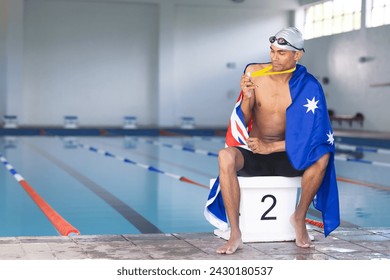 Young biracial male swimmer wrapped in an Australian flag sits poolside with a medal. Reflecting on his performance, the swimmer holds a medal at an indoor swimming facility. - Powered by Shutterstock