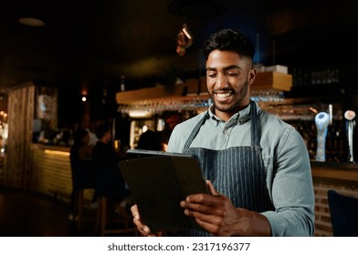 Young biracial man in apron smiling while using digital tablet while working as waiter in restaurant - Powered by Shutterstock