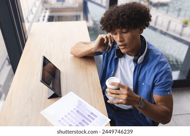 A young biracial man with curly brown hair is talking on the phone in a modern business office. He's wearing a blue shirt, holding a coffee cup, and has a tablet displaying VR graphs, unaltered. - Powered by Shutterstock